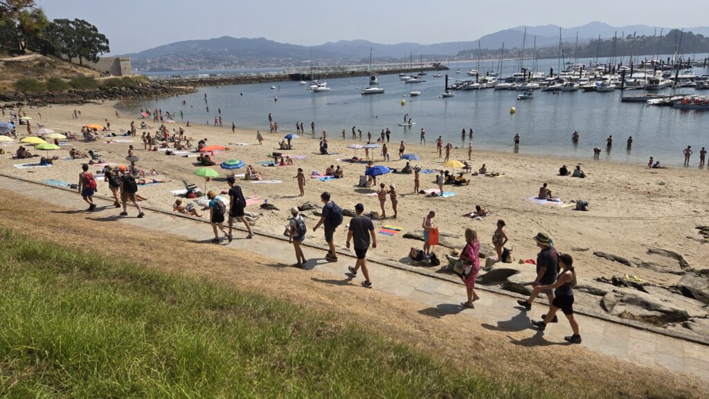 People enjoying a sunny day at the beach in Galicia with boats in the harbor. longevity in Galicia.