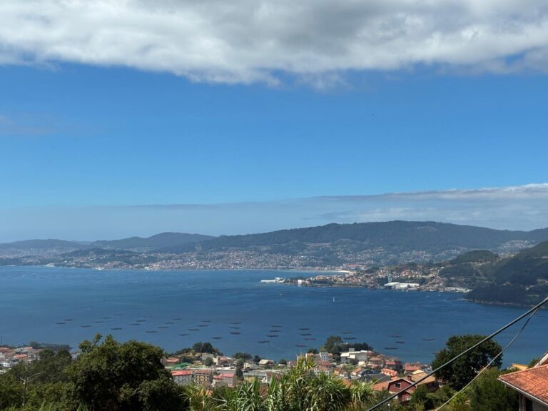 Scenic view of Galicia with a large lake, mountains, and a coastal town under a partly cloudy sky.