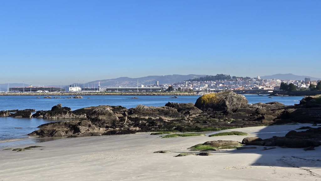 Scenic view of Galicia's coastline with rocks, sandy beach, and city skyline in the distance.