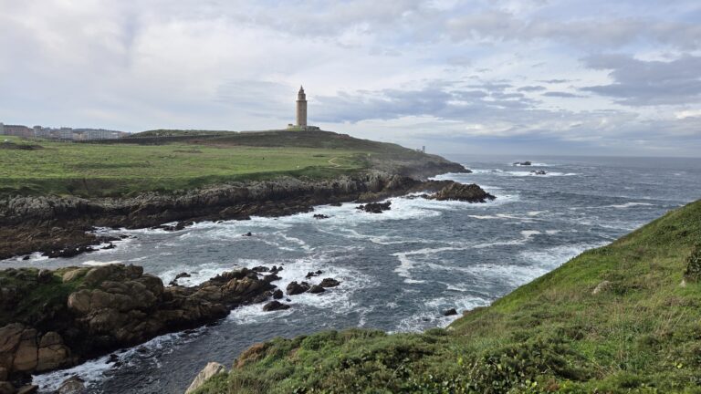 Lighthouse on rugged Galician coastline with ocean waves and cloudy sky.