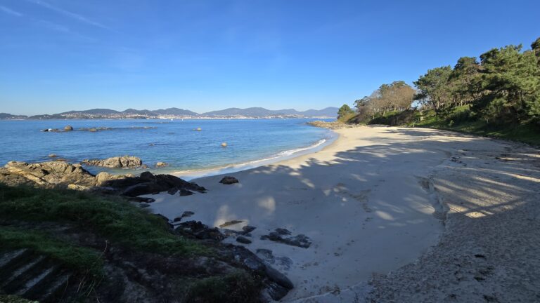 Beautiful Galician beach with clear blue water and green trees under a bright sky.