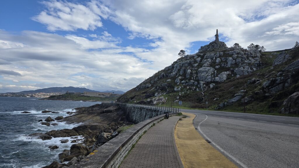 Scenic coastal road in Galicia with rocky hill and statue under cloudy sky.