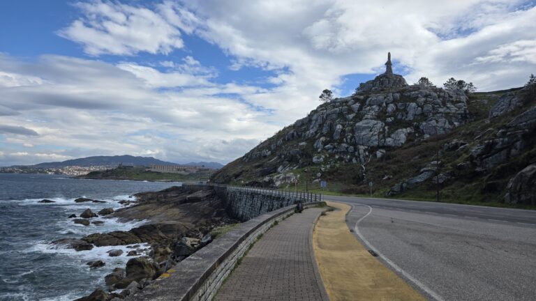 Scenic coastal road in Galicia with rocky hill and statue under cloudy sky.