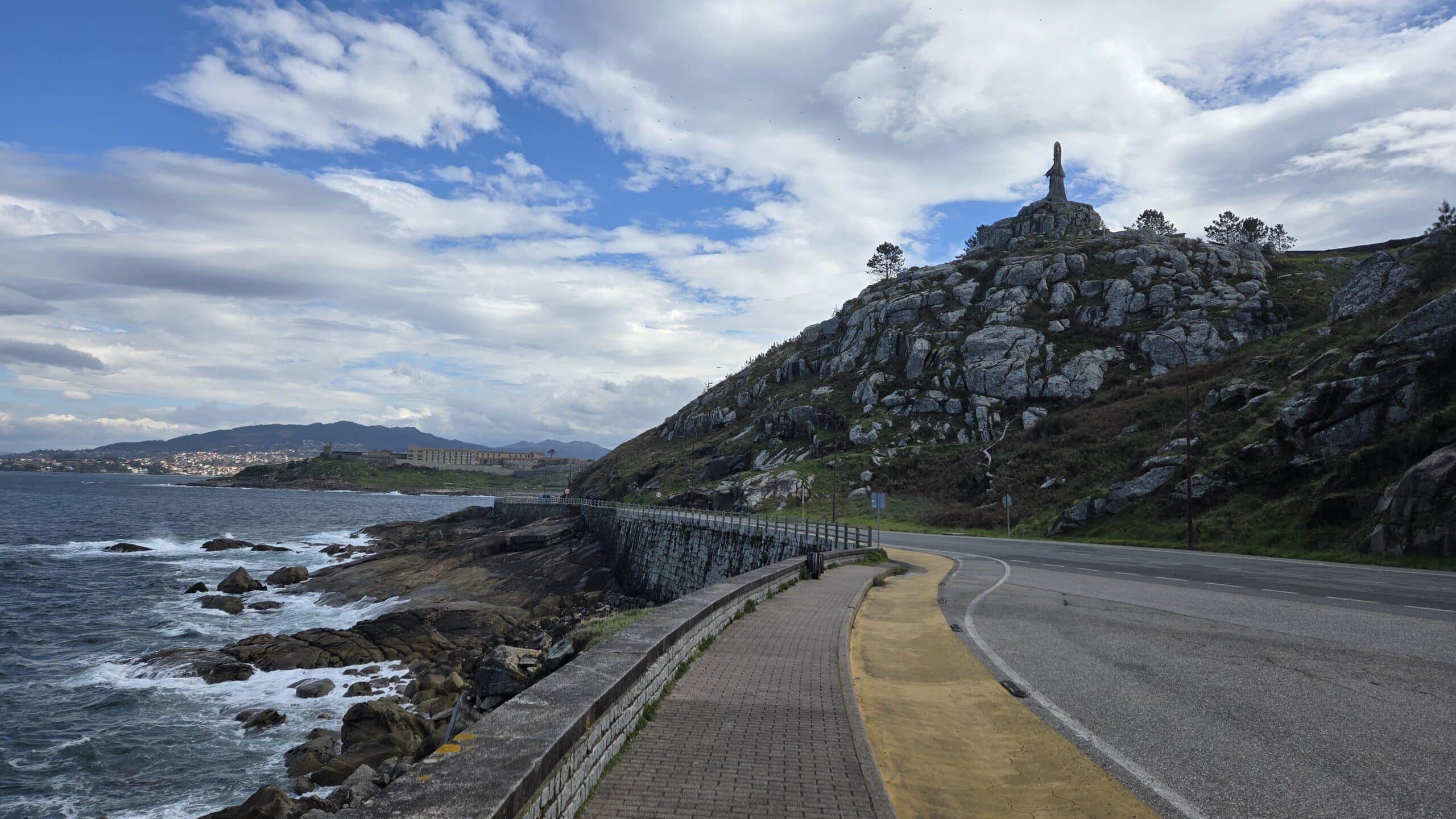 Scenic coastal road in Galicia with rocky hill and statue under cloudy sky.